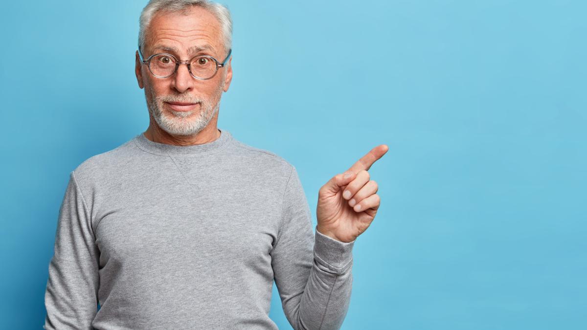 horizontal-shot-curious-grey-haired-senior-man-points-index-finger-away-blank-space-shows-place-your-advertisement-wears-casual-jumper-isolated-blue-wall_30138100