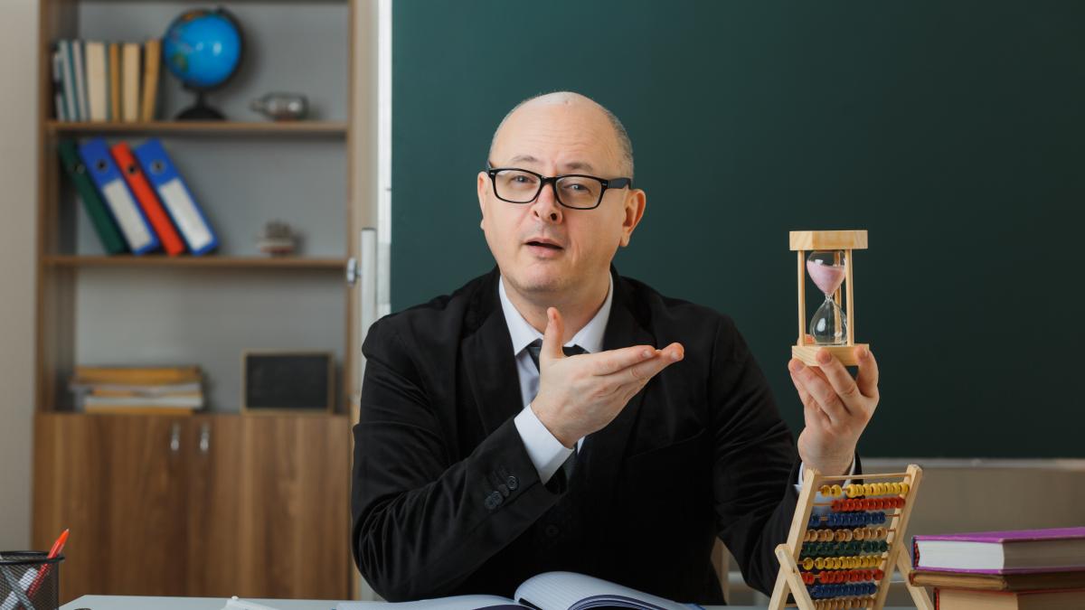 man-teacher-wearing-glasses-sitting-school-desk-front-blackboard-classroom-presenting-hour-glass-explaining-lesson-looking-confident_38123200
