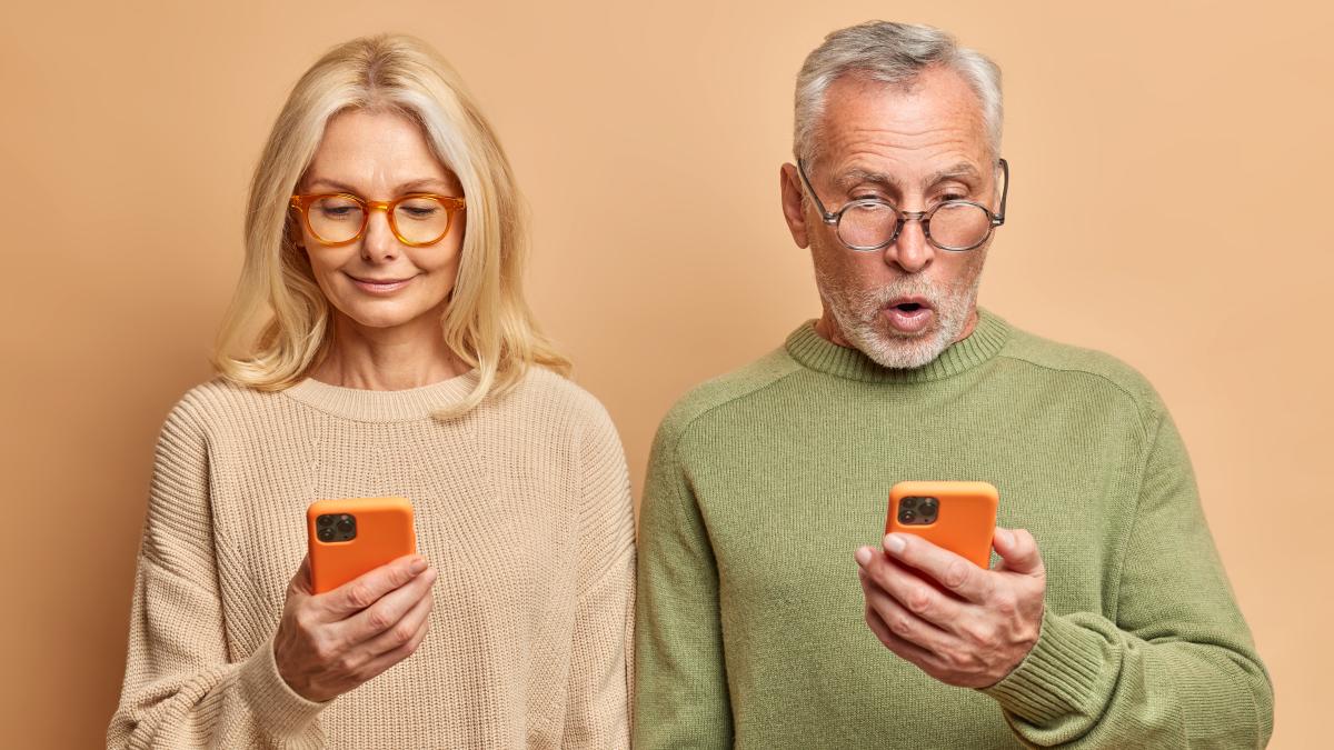 senior-couple-stand-shoulder-shoulder-use-smartphones-browsing-internet-read-media-website-dressed-casual-jumpers-isolated-brown-wall_48656700