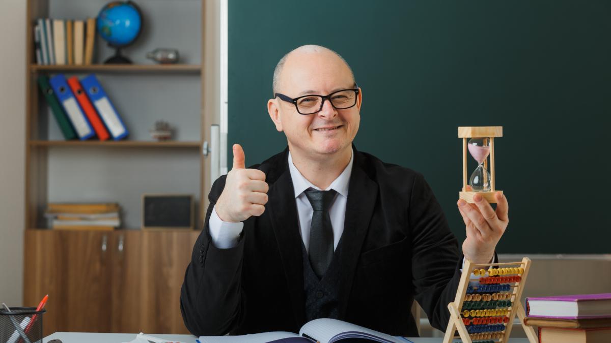 man-teacher-wearing-glasses-sitting-school-desk-front-blackboard-classroom-holding-hour-glass-explaining-lesson-showing-thumb-up-happy-pleased_77754300