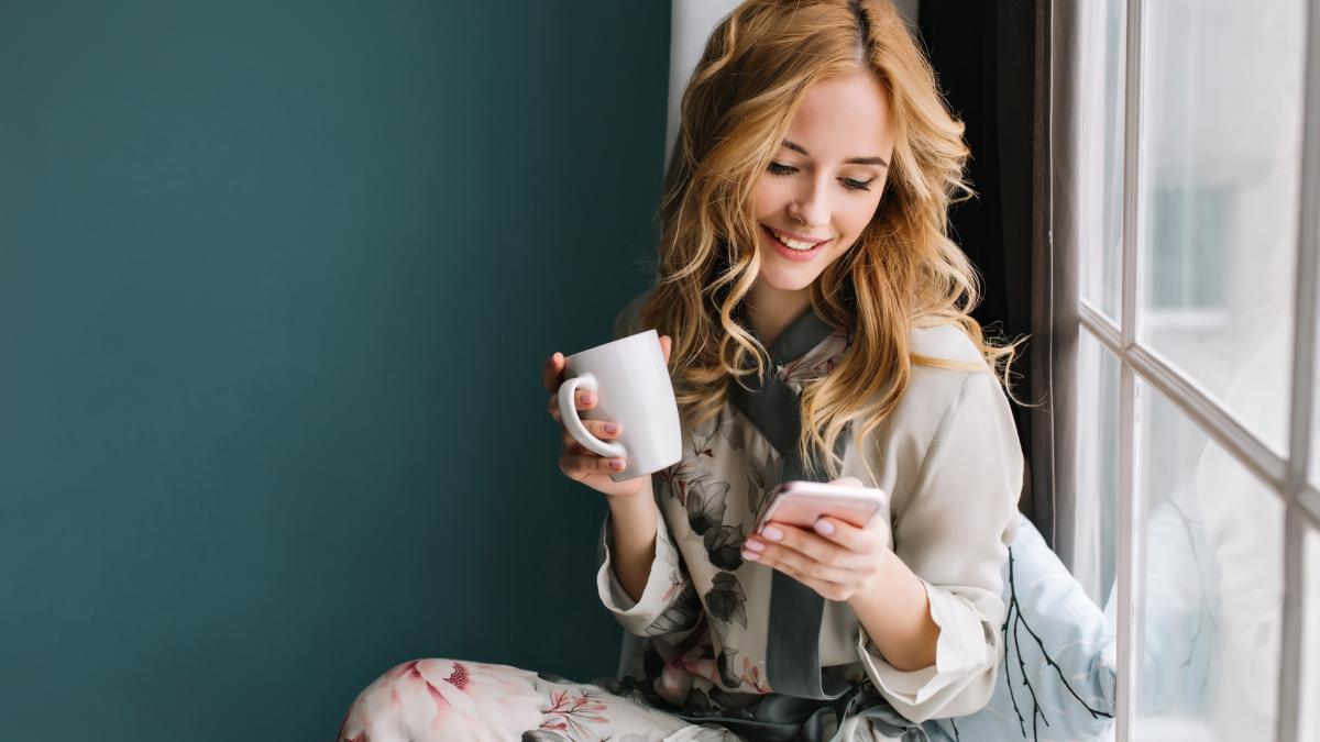pretty-blonde-girl-sitting-window-sill-with-cup-coffee-tea-smartphone-hands-she-has-long-blonde-wavy-hair-smile-looking-her-phone-wearing-beautiful-silk-pajama_61716800