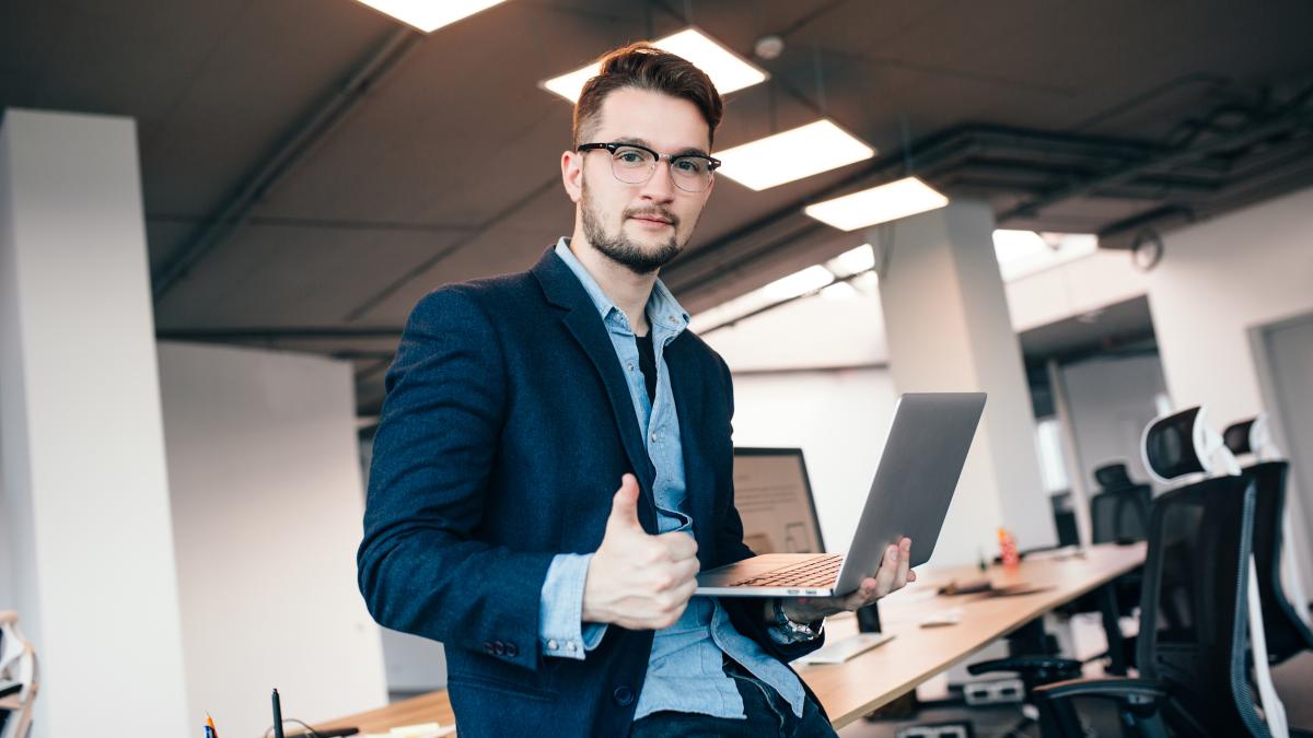 attractive-man-glassess-is-standing-near-workplace-office-he-wears-blue-shirt-dark-jacket-he-holds-laptop-shows-sign-well-looks-camera_71699500