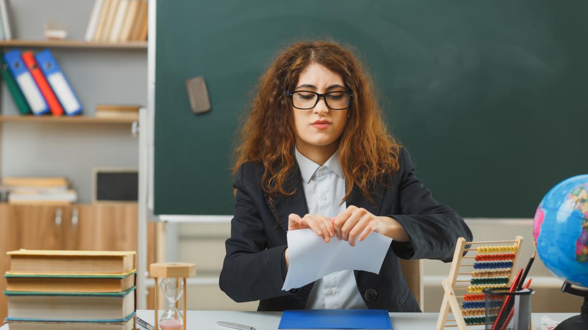 thinking-young-female-teacher-wearing-glasses-tear-paper-sitting-desk-with-school-tools-classroom_78983300