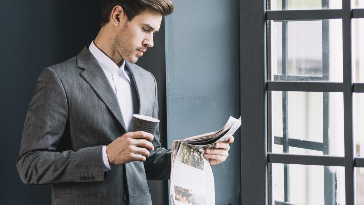young-businessman-standing-near-window-holding-cup-coffee-reading-newspaper_92457500