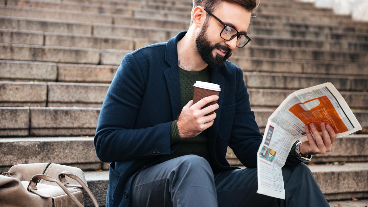 cheerful-young-bearded-man-sitting-outdoors-steps-reading-newspaper_37908000